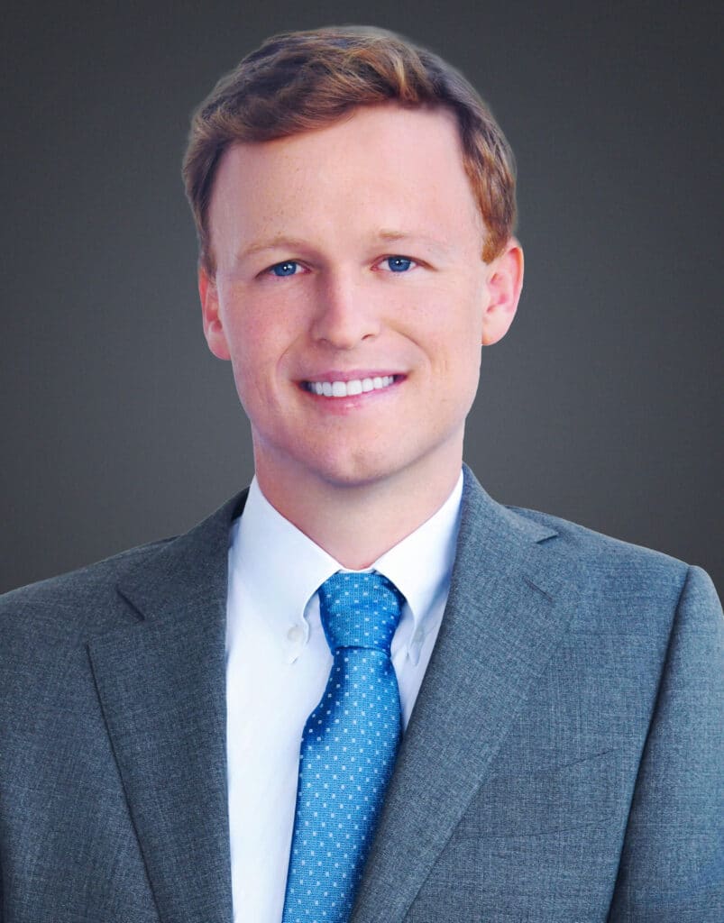 A professional headshot of Dr. Crist in a gray suit. He is wearing a white collared shirt and a blue tie with white dots, and is looking directly at the camera. The background is a solid dark gray.